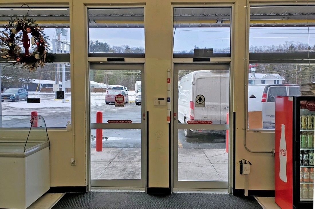 Interior view of a store entrance with glass doors. A van is parked outside. A wreath is on the left door, and a soda fridge is on the right. Overcast sky visible through the windows.