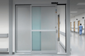 Automatic sliding glass door in a hospital hallway, with a person in scrubs walking away down the corridor—ideal for commercial doors needing reliable operation and easy access. Perfect for spaces requiring expert commercial door installation.